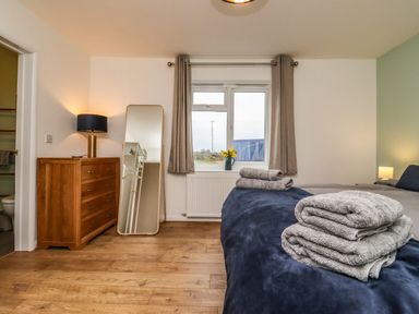A bedroom with a bed covered in blue and gray bedding and folded towels on top a wooden dresser with a lamp next to a standing mirror and a window with curtains and flowers on the sill at Harran Annex in Peopleton