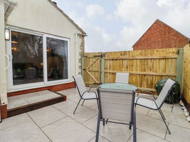 A tiled outdoor patio with a glass table and four chairs next to a wooden fence and sliding glass door at Harran Annex in Peopleton