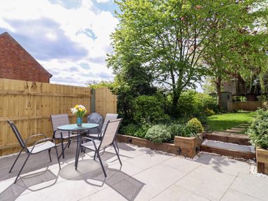 An outdoor patio with a glass table and four chairs next to a wooden fence and a garden with trees at Harran Annex in Peopleton