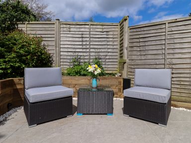 An outdoor patio with two gray cushioned chairs and a small wicker table with flowers at Harran Annex in Peopleton