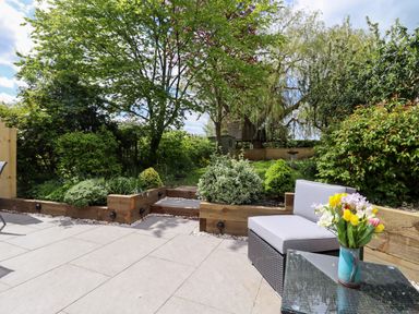 A patio area with a cushioned chair and a glass table with flowers overlooking a garden with trees and shrubs at Harran Annex in Peopleton