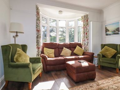 A living room with green armchairs a brown sofa and ottoman and a bay window with floral curtains at Penang in St Austell