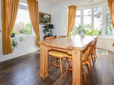 A dining room with a wooden table and chairs with yellow cushions by windows with yellow curtains and plants in Penang in St Austell