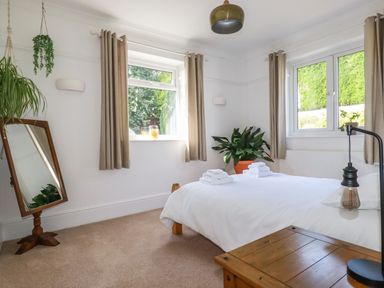 A bedroom with a bed with white bedding and folded towels two windows with beige curtains a full length mirror and green plants at Penang in St Austell