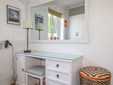 A white desk with drawers and a chair in front of a large mirror a lamp on the desk and a woven basket next to the desk at Penang in St Austell