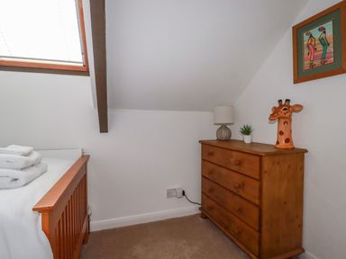 A bedroom corner with a wooden bed frame, folded towels, a wooden chest of drawers with a lamp, plant, and giraffe ornament at Penang in St Austell