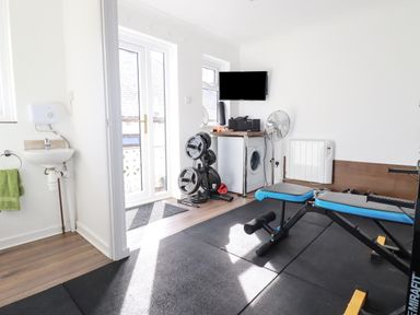 A home gym room with weightlifting bench barbell weights wall mounted tv fan and washing machine at Penang St Austell
