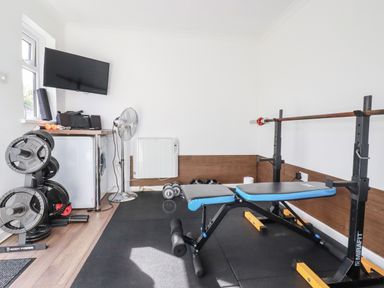 A home gym with weight plates on a rack a bench press with a barbell dumbbells a floor fan a television and a washing machine at Penang in St Austell
