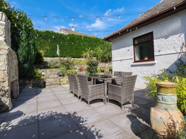An outdoor patio area with a glass table and six wicker chairs next to a white house with a window and string lights above at Penang in St Austell