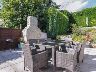 An outdoor patio area with a glass-top table surrounded by eight wicker chairs a stone fireplace string lights and green shrubs at Penang in St Austell