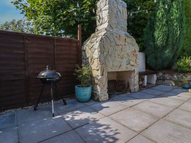An outdoor patio with a stone fireplace a black barbecue grill a blue plant pot and greenery at Penang in St Austell