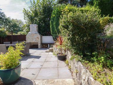 An outdoor patio with a stone fireplace a dining table and chairs surrounded by plants and bushes at Penang in St Austell