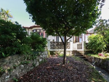 A garden path with a tree and shrubs leading to steps and a house with bay windows at Penang in St Austell