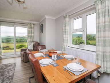 A dining area with set table and leather chairs next to windows with curtains showing green landscape at Ashdale in Bwlch-y-sarnau near Rhayader