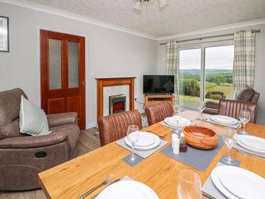 A dining area with a wooden table set with plates and glasses adjacent to a living room with armchairs, a fireplace, and a television with large windows showing a landscape at Ashdale in Bwlch-y-sarnau near Rhayader