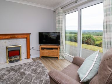 A living room with a fireplace a television on a wooden stand a chair with a cushion and sliding glass doors showing a landscape at Ashdale in Bwlch-y-sarnau near Rhayader