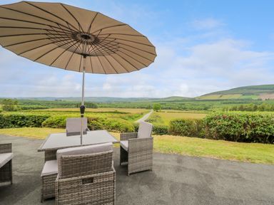 An outdoor patio with a table, chairs, and an umbrella overlooking fields and distant hills at Ashdale in Bwlch-y-sarnau near Rhayader