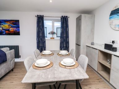 A dining area with a table set for four near a window with dark curtains and a kitchen cabinet at 2A The Anchorage in Beadnell
