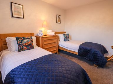 A bedroom with two single beds with blue quilts and decorative pillows with a crab design and a wooden chest of drawers with a lamp and towels at 2A The Anchorage in Beadnell