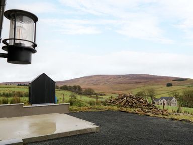 Outdoor area with a concrete slab a black shed a pile of logs and hills in the background at The Croft in Newtown-Crommelin near Cloughmills