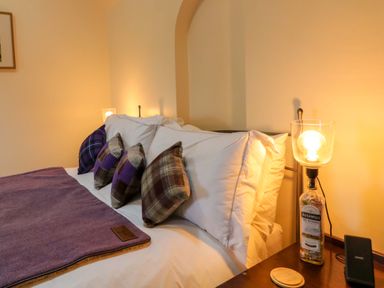 A bed with white pillows and plaid cushions next to a bedside table with a lamp made from a bottle at The Croft in Newtown-Crommelin near Cloughmills