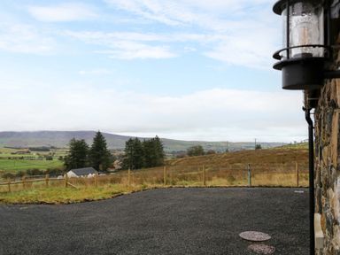 An outdoor view of a gravel driveway next to a stone wall with a light fixture and a countryside landscape with trees and hills at The Croft in Newtown-Crommelin near Cloughmills