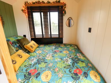 A bedroom with a floral bedspread green and yellow pillows next to a window with blinds and string lights at Herdwick Luxury Glamping Nottingham