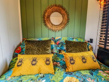 A bed with floral bedding and pillows with bee designs below a round ornate mirror on a green paneled wall at Herdwick Luxury Glamping in Nottingham