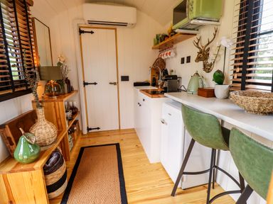 A small kitchen with white cabinets green chairs a countertop shelving and various kitchen appliances at Herdwick Luxury Glamping in Nottingham