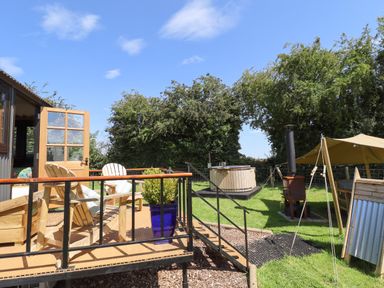 An outdoor deck with wooden chairs and a blue plant pot next to a grassy yard with a hot tub and a wood stove at Herdwick Luxury Glamping in Nottingham