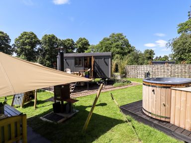 An outdoor area with a wooden hot tub a small cabin and a shaded seating area at Herdwick Luxury Glamping in Nottingham