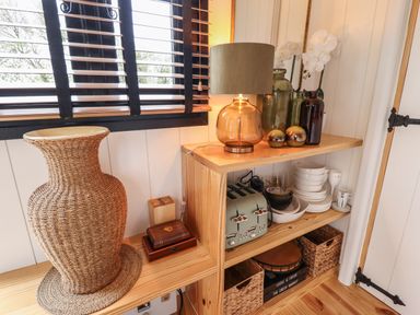A corner wooden shelf with a lamp dishes toaster vases and woven basket next to a window with blinds at Southdown Luxury Glamping in Nottingham
