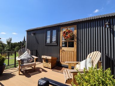 A small outdoor deck with two wooden chairs with cushions a wooden box and a door with a wreath on a black corrugated building at Southdown Luxury Glamping in Nottingham