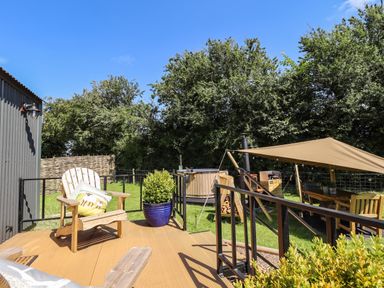 An outdoor deck with wooden chairs and a cushion near a hot tub and shaded seating area surrounded by trees at Southdown Luxury Glamping in Nottingham