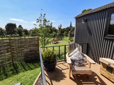 A wooden chair with cushions on a small patio next to a black corrugated building with a garden and wooden fence in the background at Southdown Luxury Glamping in Nottingham