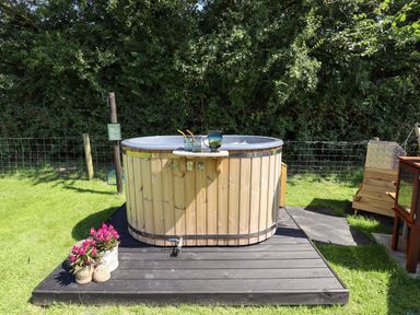 An outdoor wooden hot tub on a black platform with drinks on the edge and flowers in pots nearby at Southdown Luxury Glamping in Nottingham