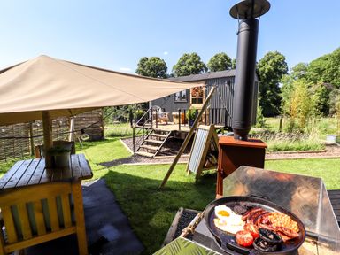 A view of an outdoor seating area with a wooden table under a canopy and a small cabin with steps in the background and a skillet cooking breakfast in Southdown Luxury Glamping Nottingham