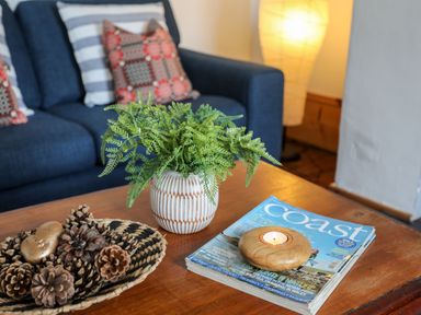 A wooden coffee table with a small plant in a white vase pine cones on a woven basket a candle on a magazine and a dark blue sofa with patterned cushions behind at East Point Lynas Lighthouse Keeper's Cottage in Llaneilian near Amlwch