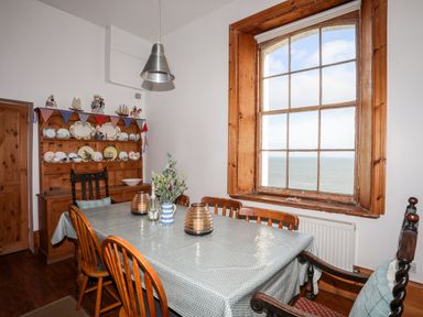 A dining room with a wooden table and chairs a hutch with plates and decorations and a large window overlooking the sea at East Point Lynas Lighthouse Keeper's Cottage in Llaneilian near Amlwch