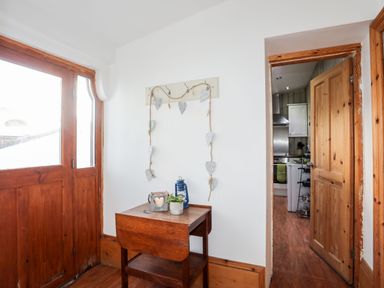 An entryway with a wooden door and small wooden table with decorative items next to an open door leading to a kitchen at East Point Lynas Lighthouse Keeper's Cottage in Llaneilian near Amlwch