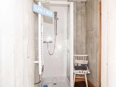 A shower cubicle next to a white chair with cushions in a wood paneled room at East Point Lynas Lighthouse Keeper's Cottage in Llaneilian near Amlwch