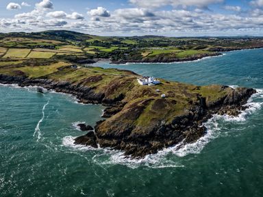 An aerial view of a rocky coastal headland with a lighthouse and buildings surrounded by the sea and fields at East Point Lynas Lighthouse Keeper's Cottage Llaneilian near Amlwch