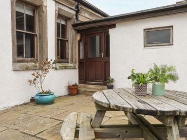 An outdoor patio area with potted plants wooden picnic table and bench in front of building with windows and a wooden door at West Point Lynas Lighthouse Keeper's Cottage Llaneilian near Amlwch