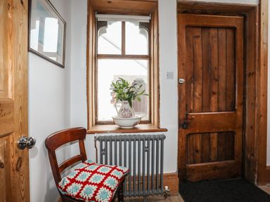A wooden chair with a crocheted cushion next to a window with a vase and bowl on the sill near a wooden door and radiator at West Point Lynas Lighthouse Keeper's Cottage in Llaneilian near Amlwch