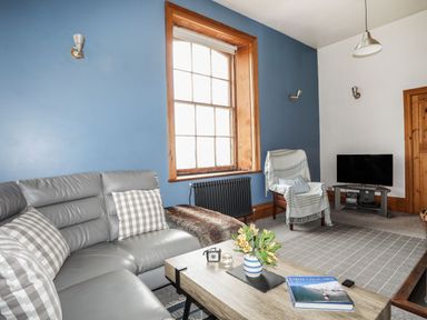 A living room with a gray sofa and checkered pillows a wooden coffee table with flowers and a book a chair with a blanket a TV on a stand blue and white walls with a large window at West Point Lynas Lighthouse Keeper's Cottage in Llaneilian near Amlwch