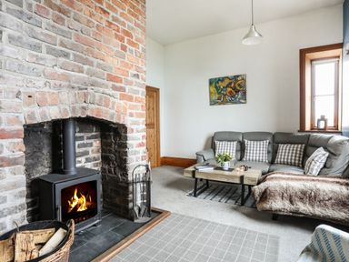 A living room with a brick fireplace and wood burning stove a gray sofa with checked cushions a wooden coffee table and a window at West Point Lynas Lighthouse Keeper's Cottage in Llaneilian near Amlwch