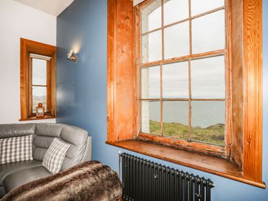 A corner with a gray sofa and checkered pillows beside two wooden framed windows overlooking water at West Point Lynas Lighthouse Keeper's Cottage in Llaneilian near Amlwch