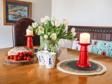 A wooden dining table with a vase of white flowers two red candles two white mugs bread and cherry tomatoes at West Point Lynas Lighthouse Keeper's Cottage in Llaneilian near Amlwch