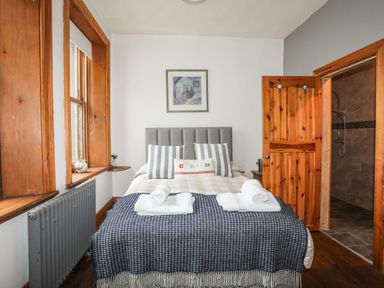A bedroom with a bed, pillows, folded towels, window, radiator, wooden door, and an attached bathroom at West Point Lynas Lighthouse Keeper's Cottage Llaneilian near Amlwch