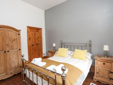 A bedroom with a metal bed with yellow pillows and blanket two wooden bedside tables with lamps a wooden wardrobe and wooden door at West Point Lynas Lighthouse Keeper's Cottage Llaneilian near Amlwch